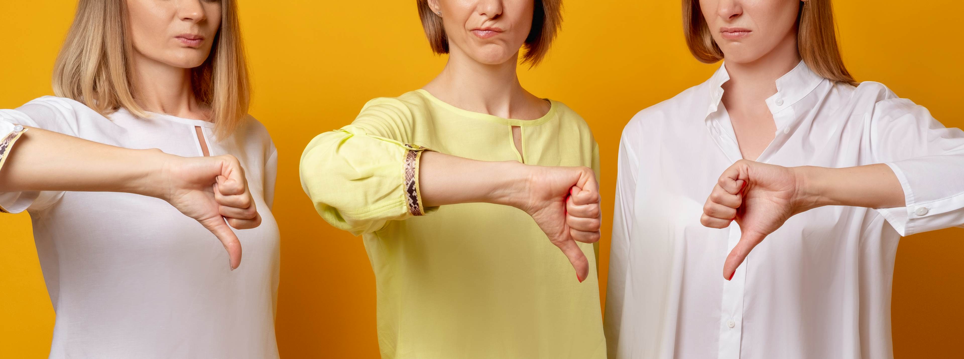 Three women standing against a bright orange background, each giving a thumbs-down gesture with serious facial expressions, symbolizing disapproval or dissatisfaction.
