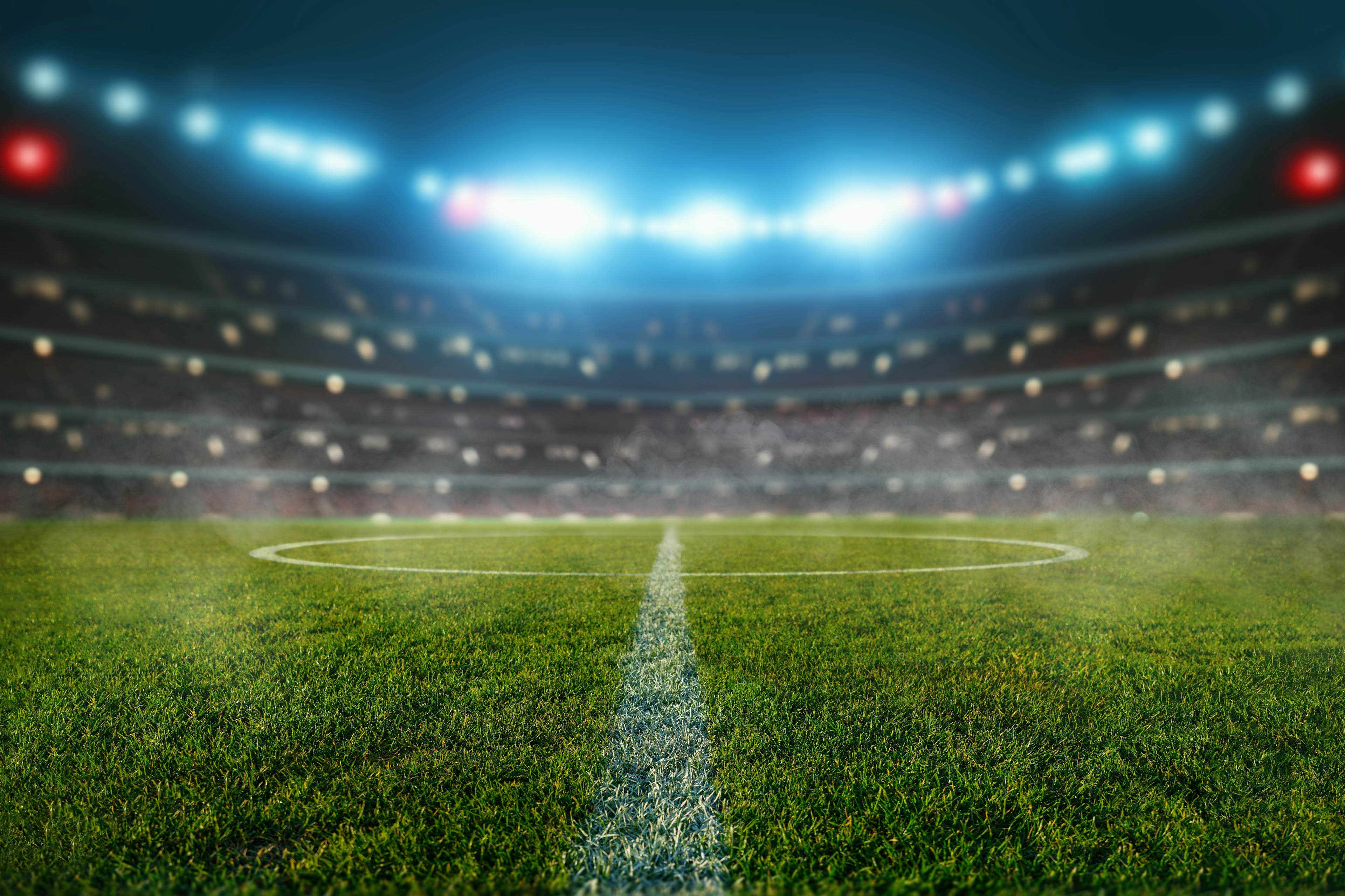 Empty soccer stadium with bright lights, green pitch, and visible midfield line under night sky.