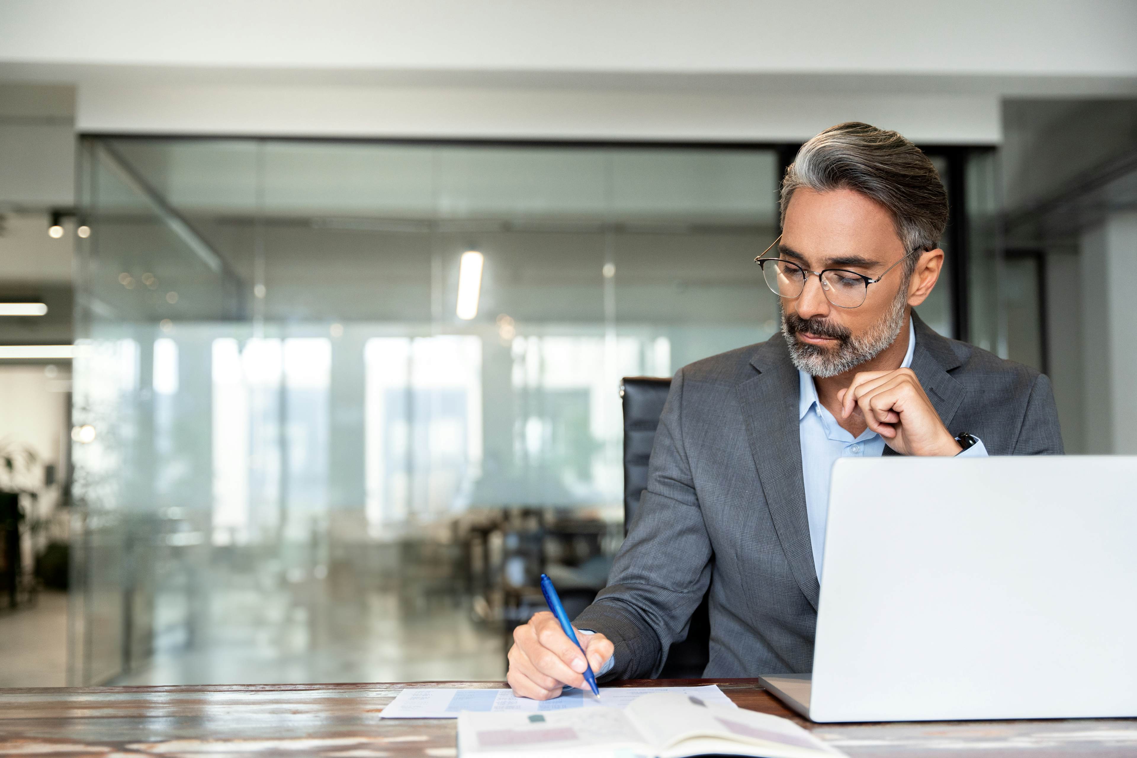 Businessman in a suit working at a desk with a laptop, writing in a notebook in a modern glass-walled office.
